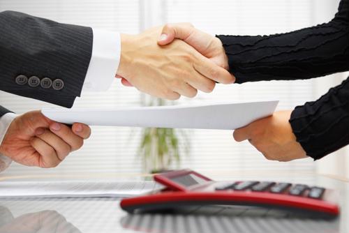 Business people shaking hands while exchanging documents above a desk.